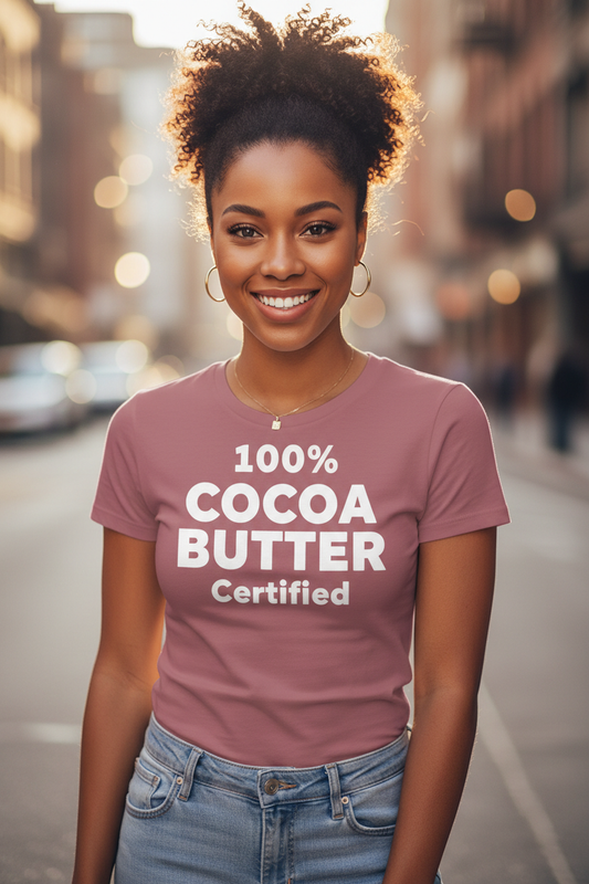 A photorealistic lifestyle mockup of a smiling African American woman in her late 20s to early 30s standing outdoors on a softly blurred city street. She has warm brown skin, bright expressive eyes, and natural curly hair styled in a high ponytail. She is wearing a fitted mauve T-shirt with bold white text that reads “100% COCOA BUTTER Certified” centered on the chest. The shirt has a feminine, flattering fit. She pairs the tee with light blue jeans and minimal gold jewelry, including hoop earrings a
